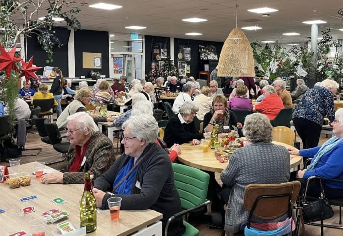 Een grote groep ouderen zit samen in een gemeenschappelijke ruimte, gezellig pratend en spelend aan tafels met decoratie en drankjes.