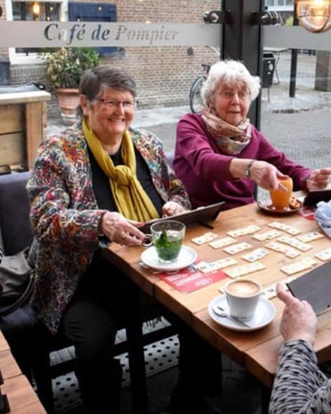 Twee oudere vrouwen zitten op een terras en spelen Rummikub, met thee en koffie op tafel. Ze lachen en genieten van het moment in een gezellig café.